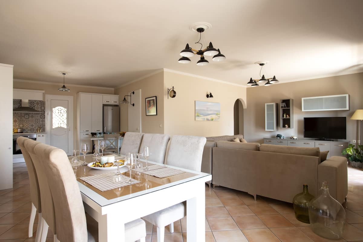 A modern living room with a dining area, a sofa, a TV, and a kitchen in the background.
