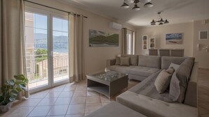 Un salon spacieux, doté d’un grand canapé, d’une table basse en verre et offrant une vue sur un balcon donnant sur un paysage pittoresque.
