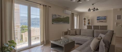 A spacious living room with a large sofa, a glass coffee table, and a view of a balcony with a scenic landscape.