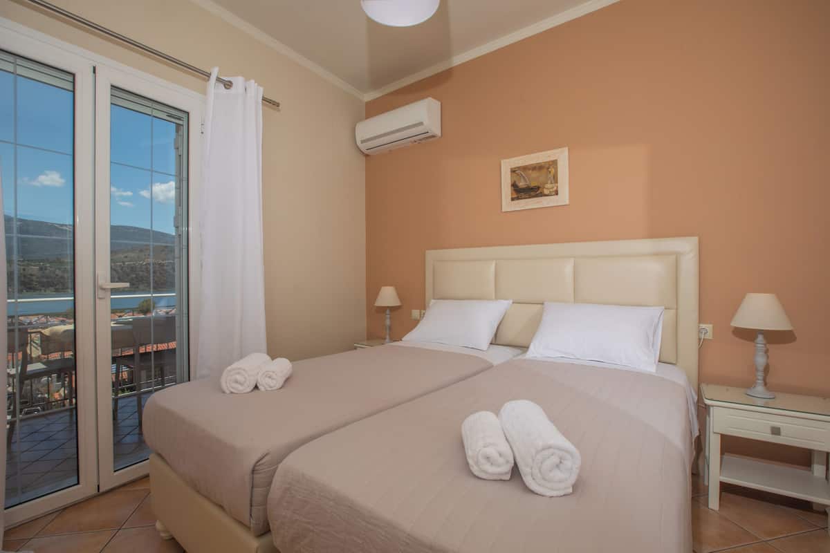 A hotel room with two beds, a balcony with a view, an air conditioning unit, and a small table with a lamp.
