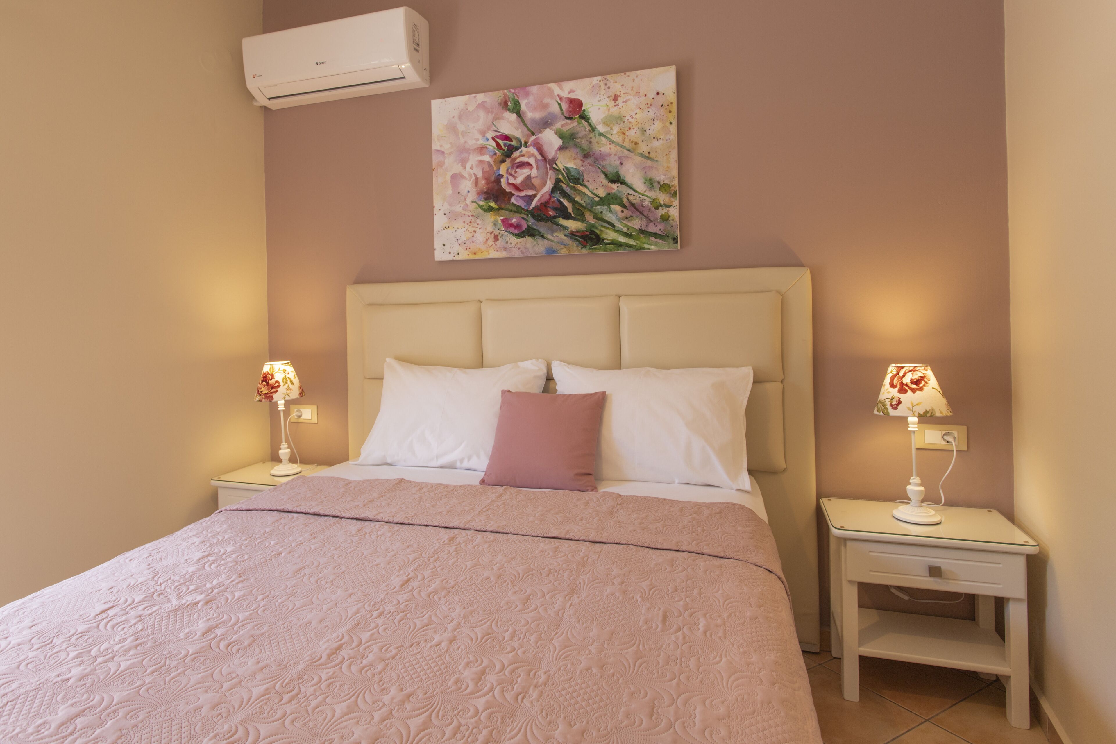 Une chambre à coucher comprenant un lit, deux tables de chevet, un climatiseur fixé au mur et un tableau représentant des motifs floraux.