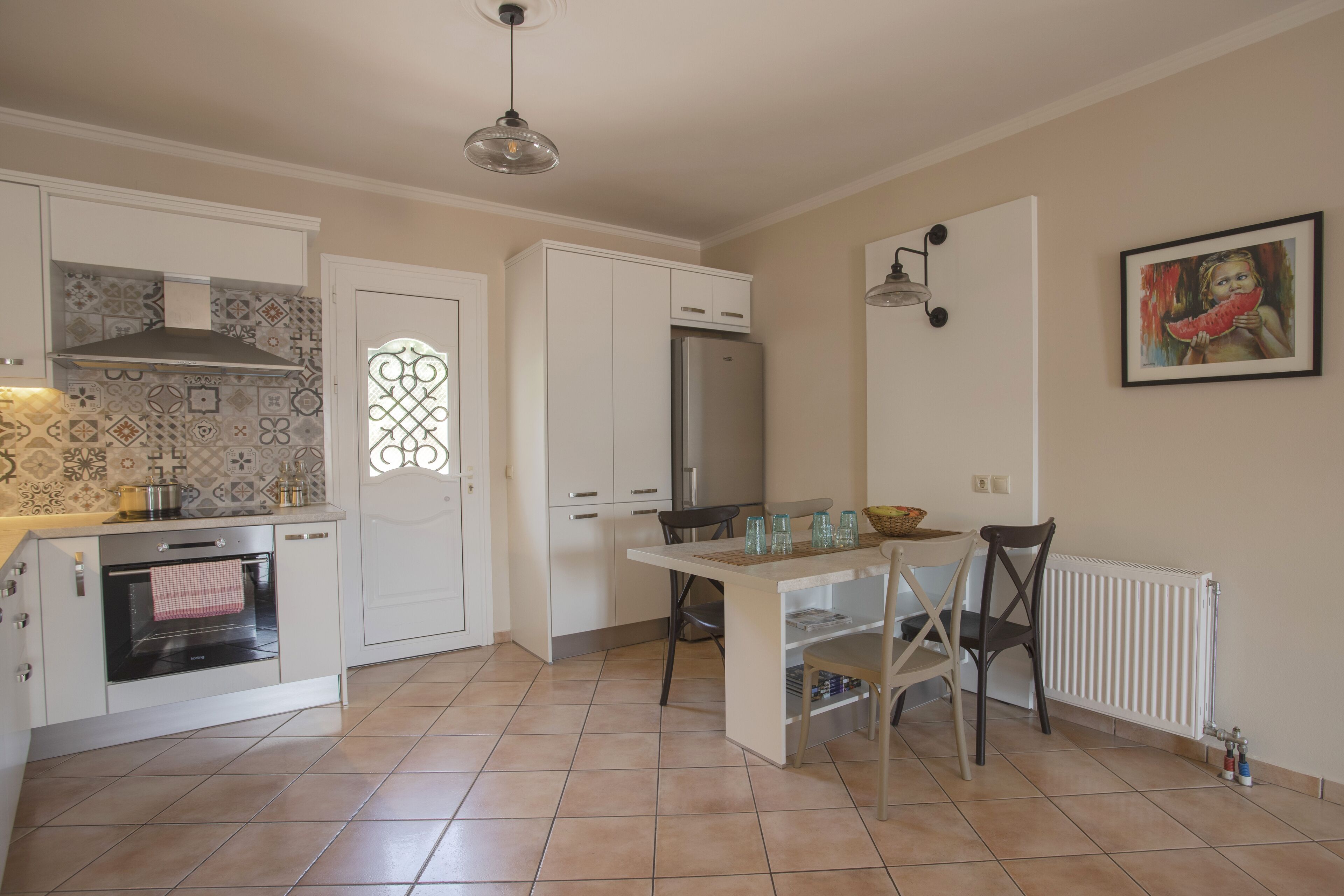 Una cocina con gabinetes blancos, horno empotrado, refrigerador, mesa de comedor con sillas y una lámpara colgante.