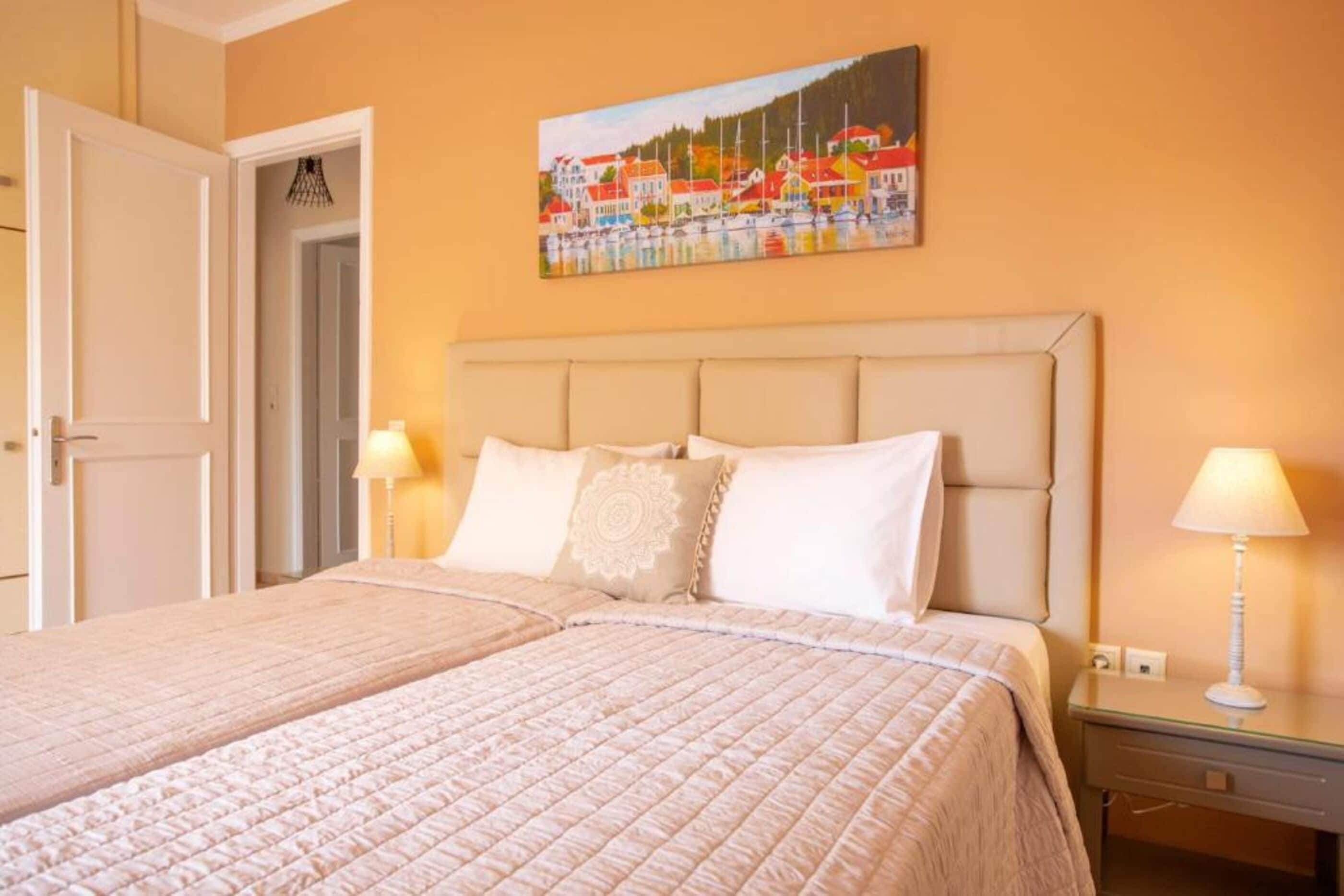 Hotelkamer met een groot bed, hoofdeinde, twee bedlampen en een schilderij van een kustplaats aan de muur.