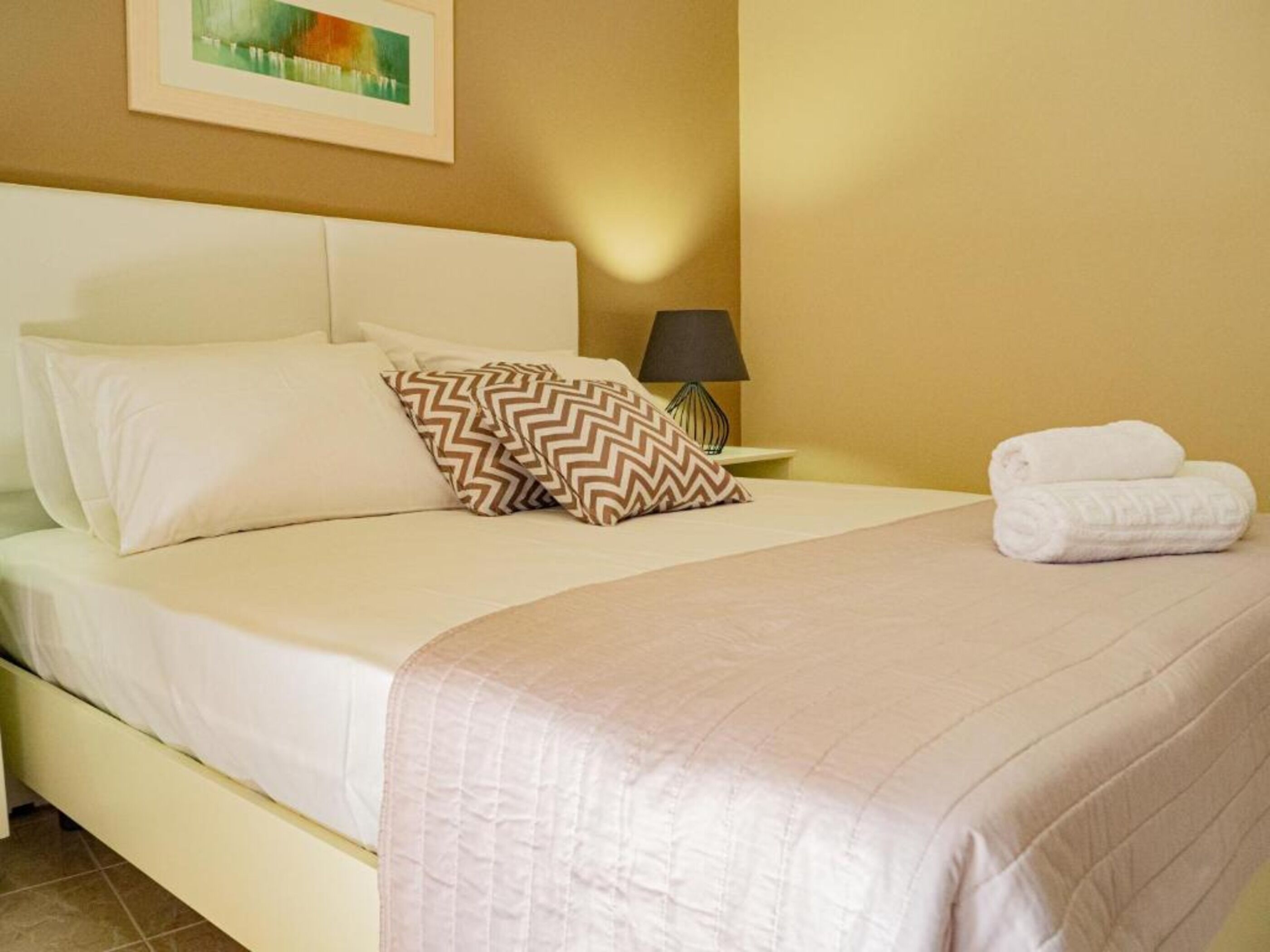 A neatly made bed with white bedding and two patterned pillows, a folded towel on the bed, and a lamp on a bedside table.
