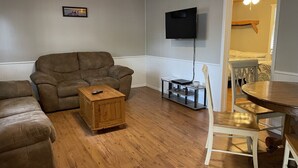 Una sala de estar con un sofá de cuero marrón, una mesa de centro de madera, un televisor montado en la pared y una mesa de comedor con sillas.