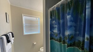 Ein Badezimmer mit Toilette, Handtuchhalter mit Handtüchern, einem Fenster mit Jalousien und einem Duschvorhang mit tropischem Muster.