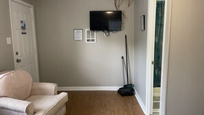 A bedroom with a bed, a chair, a television mounted on the wall, a broom, and a door leading to another room.