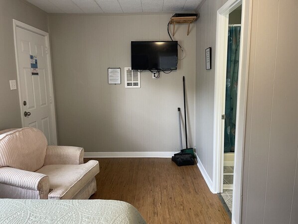 A bedroom with a bed, a chair, a television mounted on the wall, a broom, and a door leading to another room. - Quaint cabin at Little Mineral Marina: Cabin 4 (Pottsboro)