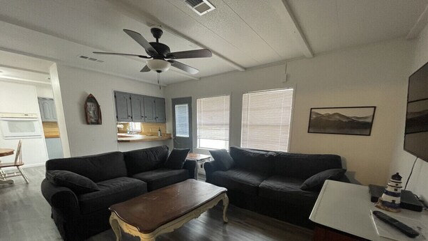 A living room with two black sofas, a wooden coffee table, a ceiling fan, and a kitchen area in the background.