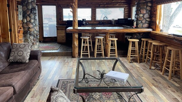 A rustic interior with a stone pillar, a brown leather sofa, a glass coffee table, wooden stools, and a kitchen area with a bar.