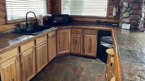 A wooden cabin kitchen with stone flooring, wooden cabinets, a sink, and a stone wall.
