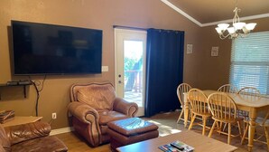A living room with a brown leather sofa, a wooden coffee table, a television mounted on the wall, a dining table with chairs, and a chandelier. - Lovely Lake House with Marina and Sunset Views (Pottsboro)