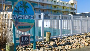 Schild einer privaten Wohnanlage, Maresol Condominium, mit Palmen und Strandmotiv, umgeben von einem weißen Zaun. Im Hintergrund ist ein mehrstöckiges Gebäude zu sehen.