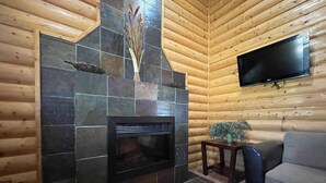 Interior de una cabaña de troncos con chimenea de piedra, un televisor de pantalla plana montado en la pared y un sofá gris.