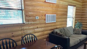 Interior de una cabaña de madera con una mesa de comedor, sillas, un sofá y dos ventanas con persianas.