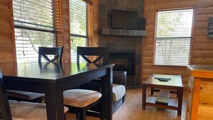Interior de una cabaña de madera con una mesa de comedor, sillas, chimenea y un televisor montado en la pared.