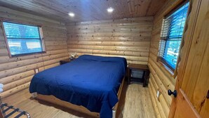 Un dormitorio tipo cabaña de troncos con cama, dos ventanas y una puerta de madera.