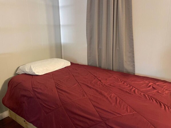 A single bed with a red bedspread and a white pillow in a room with white walls and a gray curtain.