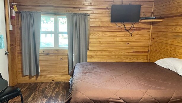 A wooden cabin room with a bed, a chair, a TV mounted on the wall, and a window with curtains. - Cozy Cedar Cabin at Mill Creek Marina: Cabin 3 (Pottsboro)