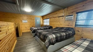 A cabin-style room with two beds, wooden walls, a ceiling fan, and a window with blinds.