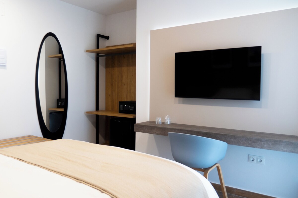 A hotel room with a bed, a desk with a chair, a flat-screen TV, a mirror, and built-in shelves.