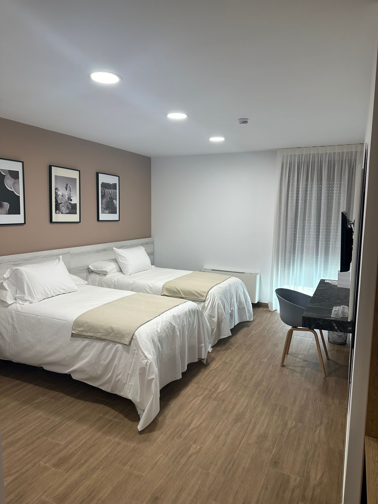 Habitación de hotel con dos camas, un escritorio y una silla. Hay luces en el techo y una ventana con cortinas.