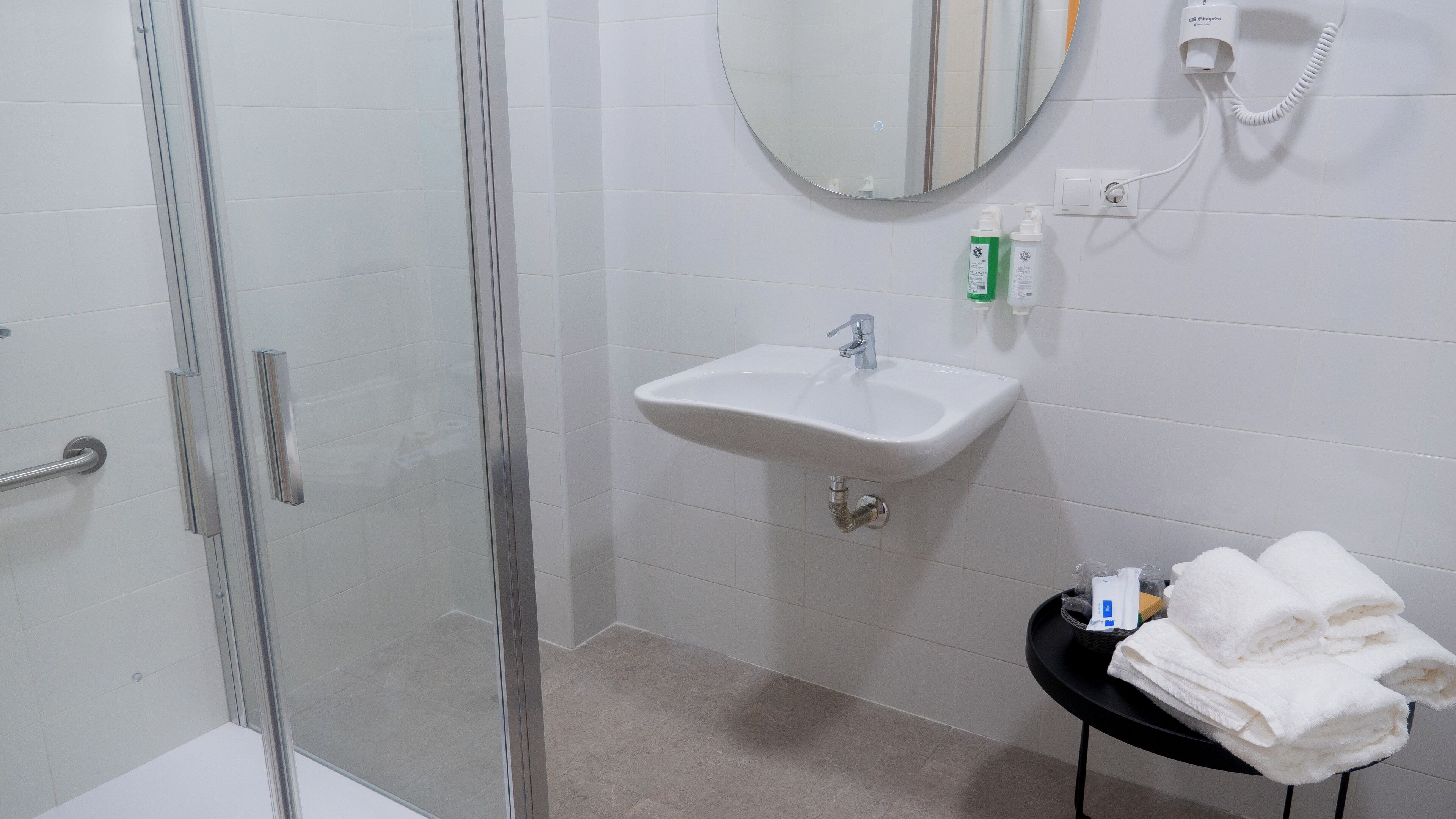 A bathroom with a glass shower enclosure, a sink with a mirror above it, and a small table with towels.