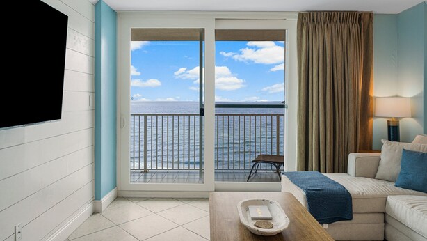 Habitación de hotel con un amplio ventanal corredero que da a un balcón con vistas al mar. Cuenta con un televisor de pantalla plana en la pared, un sofá y una mesita con bandeja.