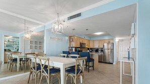 Una cocina amplia con armarios de madera, un comedor con mesa blanca y sillas azules, y una lámpara de techo moderna.