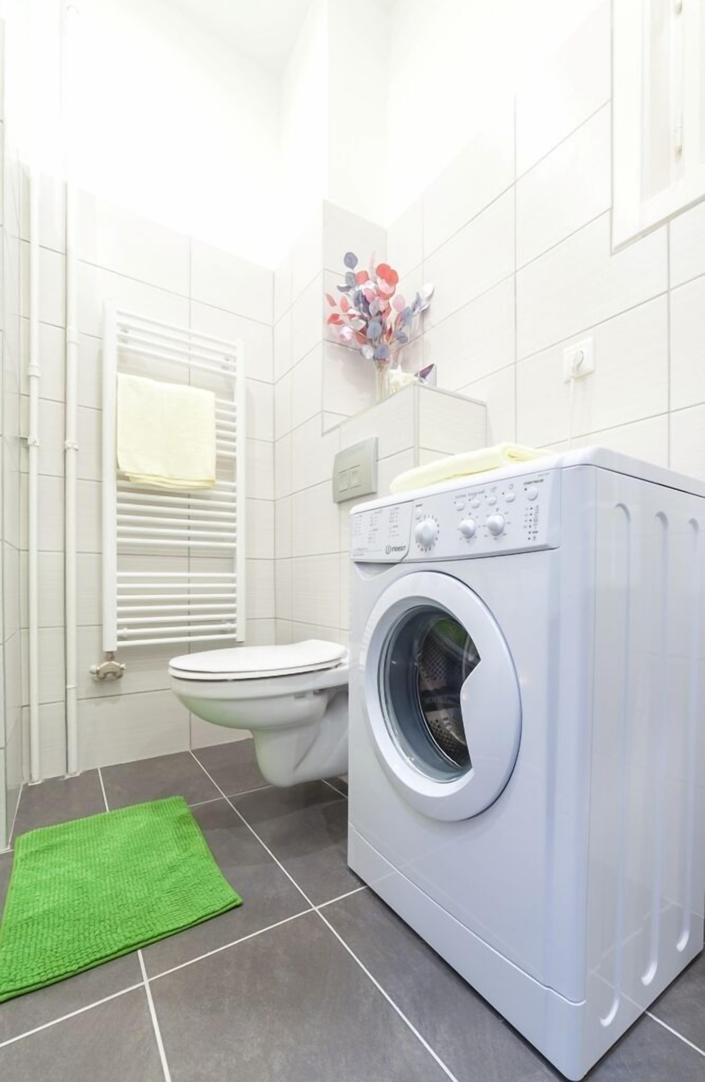 A compact bathroom with a white tiled wall, a white toilet, a washing machine, and a green mat on the floor.