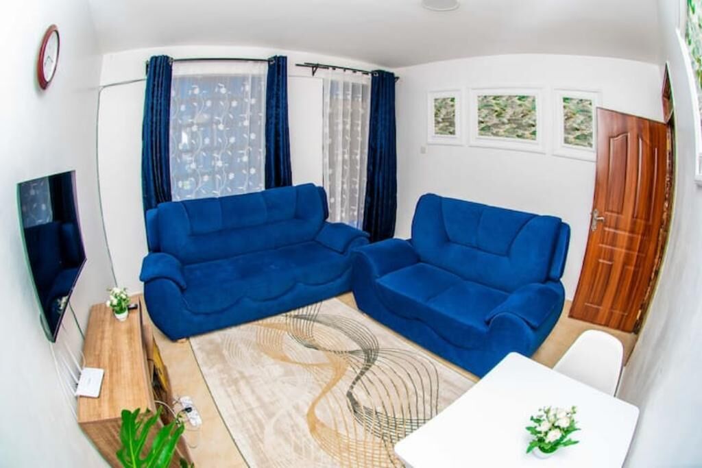 Een knus leefruimte met een blauwe zithoek, een klein houten bijzettafeltje, een witte tafel met een vaas, en een aan de muur bevestigde televisie.