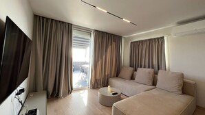 Una habitación de hotel moderna con una televisión de pantalla plana, un sofá, una mesa de centro redonda y un ventanal corredero que da a un espacio exterior.