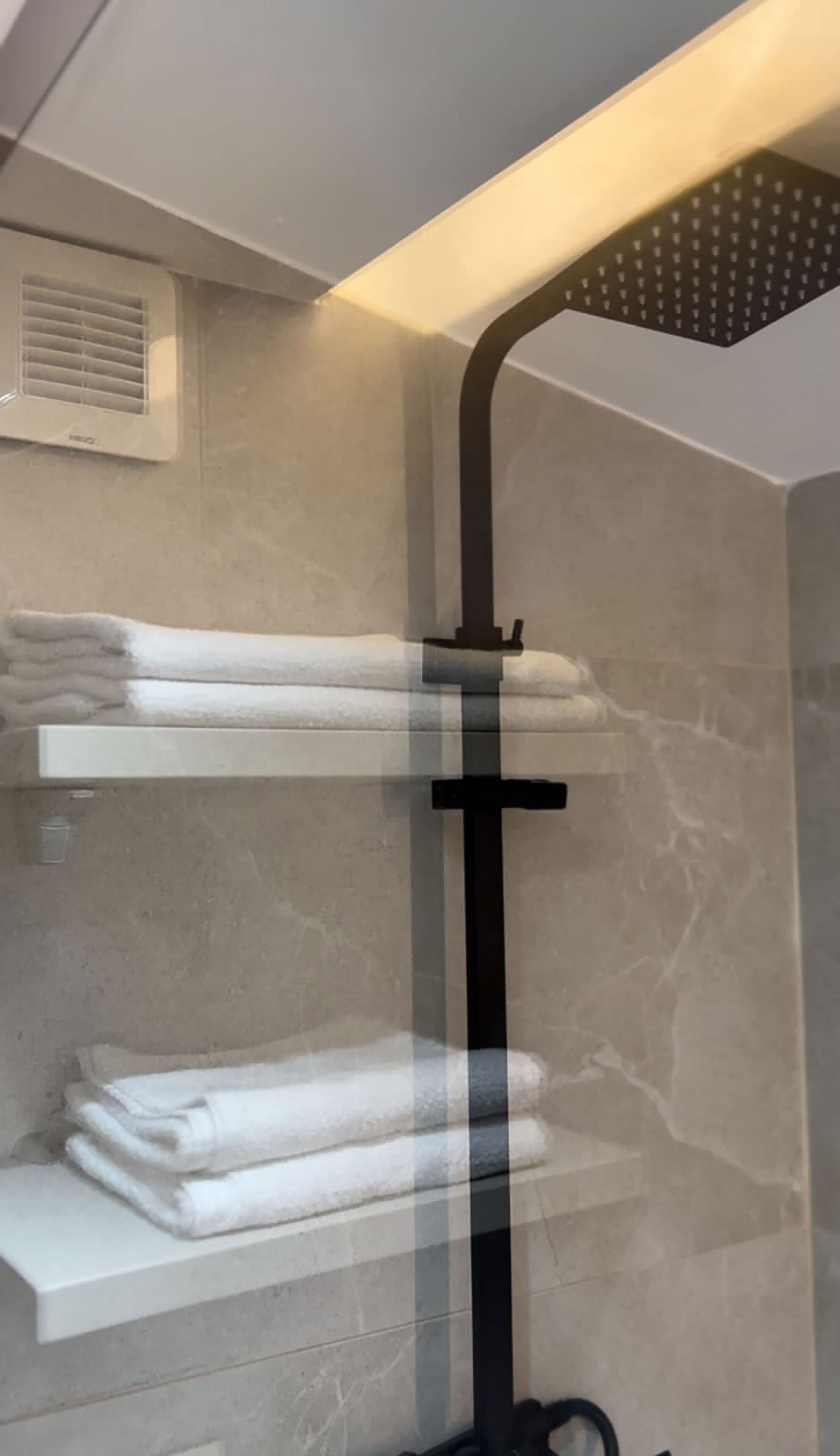 A bathroom with a glass-enclosed shower, marble walls, and shelves holding folded towels.