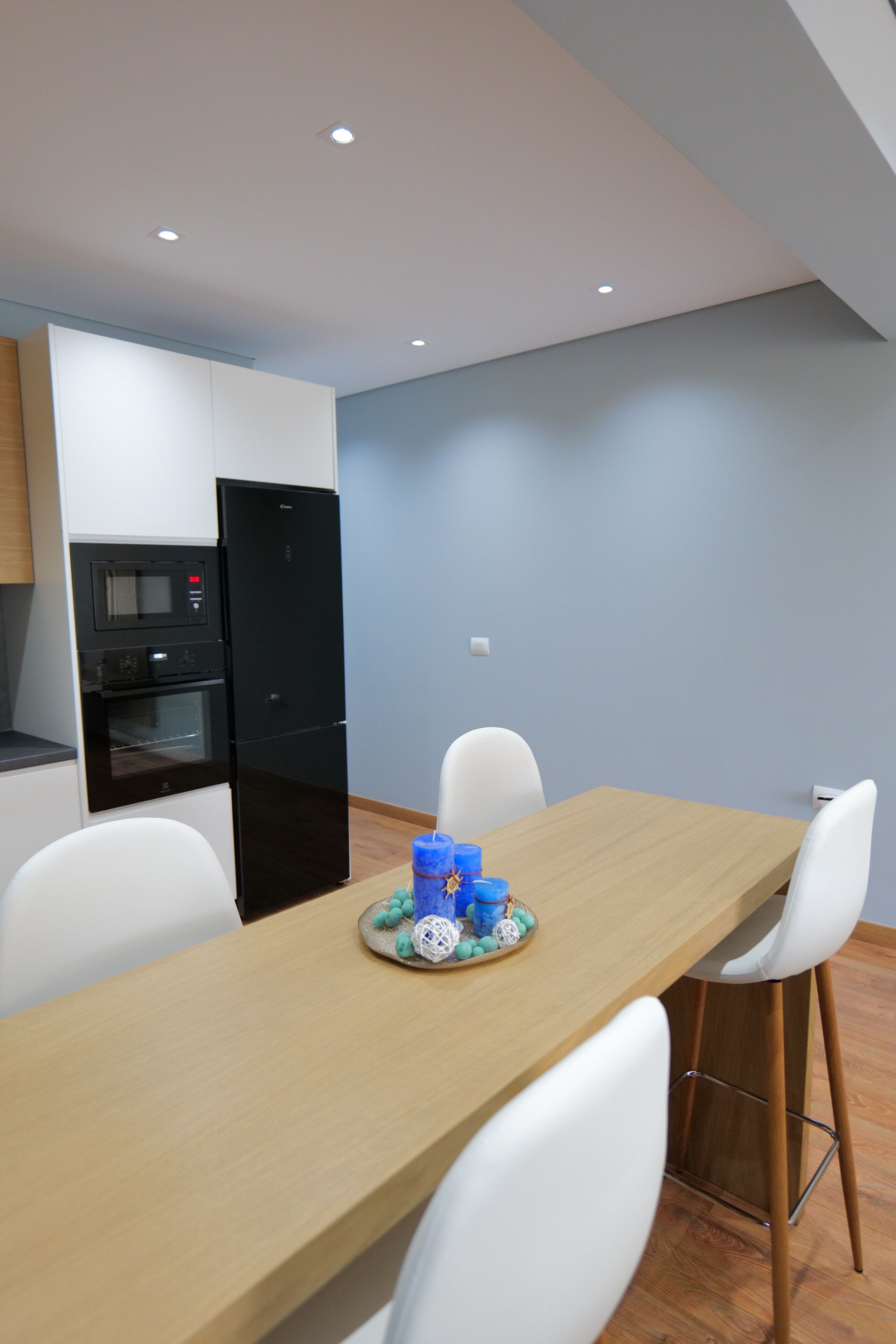 A modern kitchen with a built-in microwave and oven, a black refrigerator, and a wooden dining table with white chairs.
