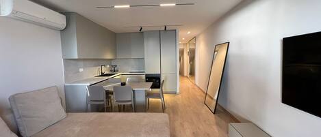 Uma sala de estar moderna com um sofá, uma área de jantar com cadeiras, uma cozinha com armários e uma televisão grande fixada na parede.