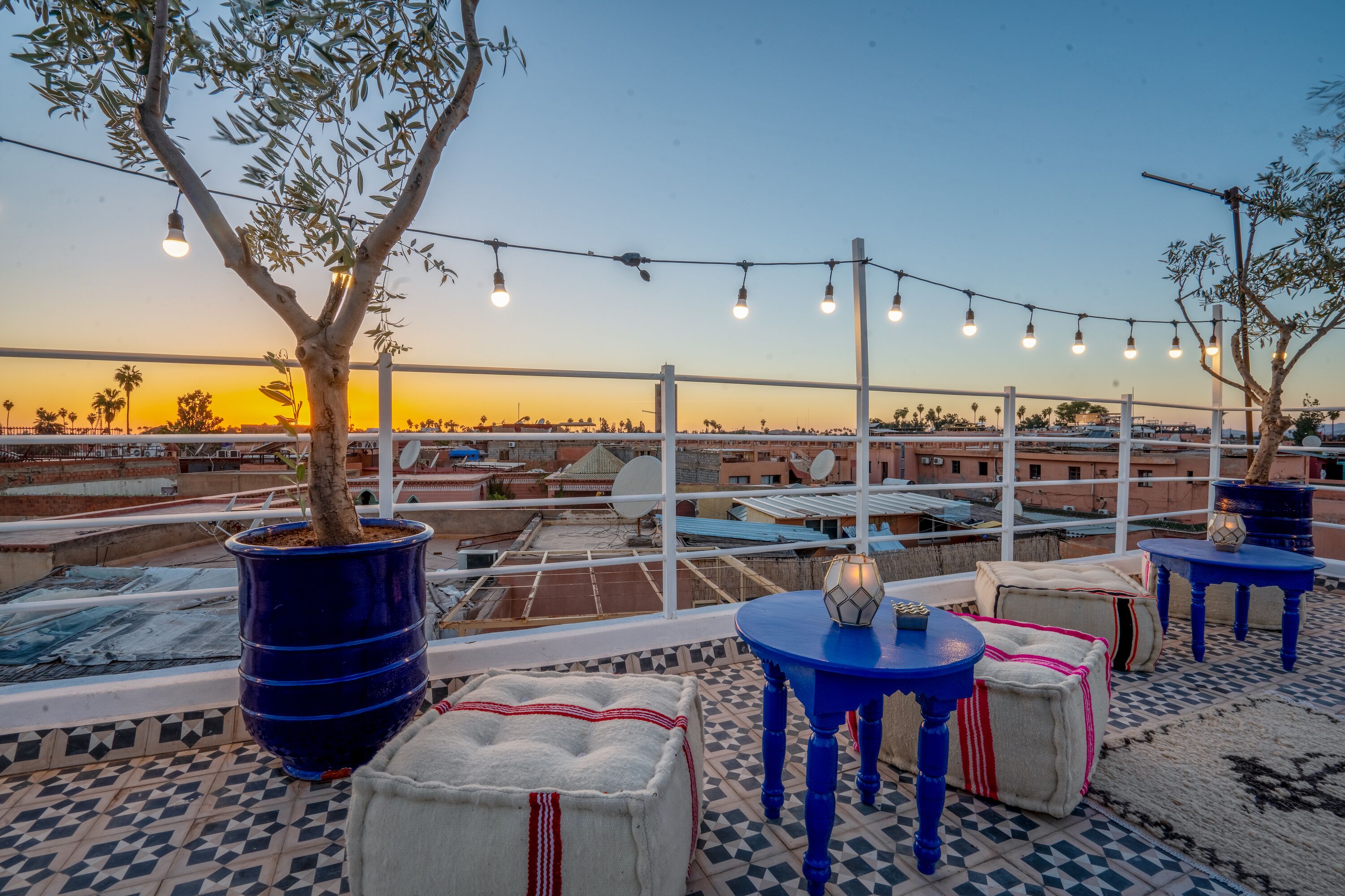 A rooftop terrace with potted plants, tables, and string lights against a sunset backdrop.