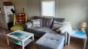 A living room with a grey sofa, a small white table, a grey ottoman, a window with blinds, a lamp on a side table, and a refrigerator. - Cozy Two Bedroom North Shore Cottage (GRAND TRACADIE)