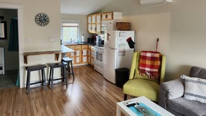A modern kitchen with wooden cabinets, a built-in oven, and a refrigerator. There is a dining area with a wooden table and two stools. - Cozy Two Bedroom North Shore Cottage (GRAND TRACADIE)