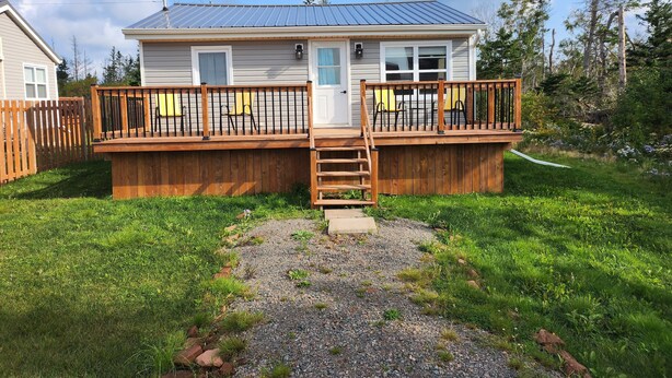 A two-story house with a deck, a front door, and a small front yard. - Cozy Two Bedroom North Shore Cottage (GRAND TRACADIE)