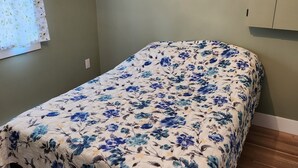 A bedroom with a floral bedspread, a window with a floral curtain, and a wooden floor. - Cozy Two Bedroom North Shore Cottage (GRAND TRACADIE)