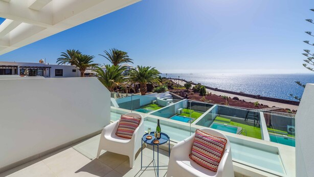 Ein Balkon mit zwei weißen Stühlen, einem kleinen Tisch mit Flasche und Gläsern sowie einem überdachten Poolbereich mit Meerblick.