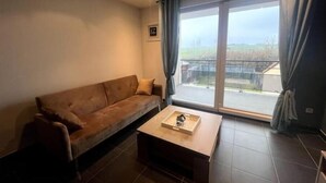 Sala de estar com um sofá marrom, uma mesa de centro e uma janela grande com cortinas.