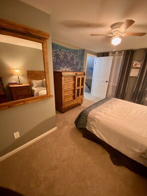 Um quarto com cama, penteadeira, espelho, ventoinha no teto e porta que dá para outro cômodo.