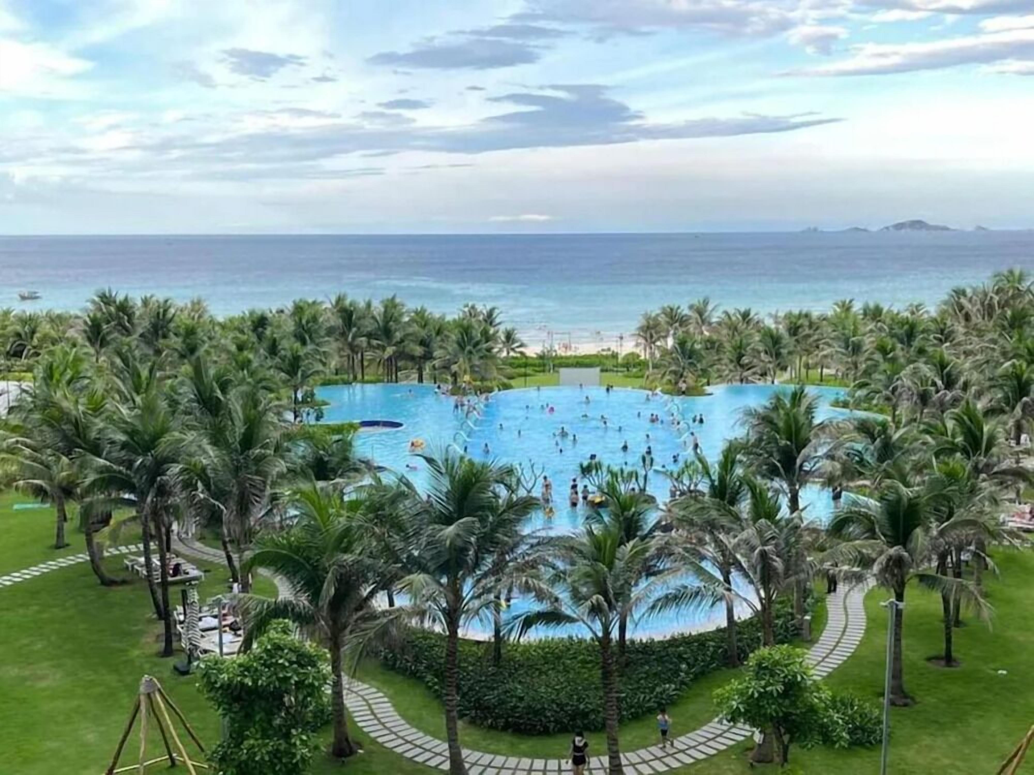 A large outdoor swimming pool surrounded by palm trees, with a view of the ocean and a beach in the background.