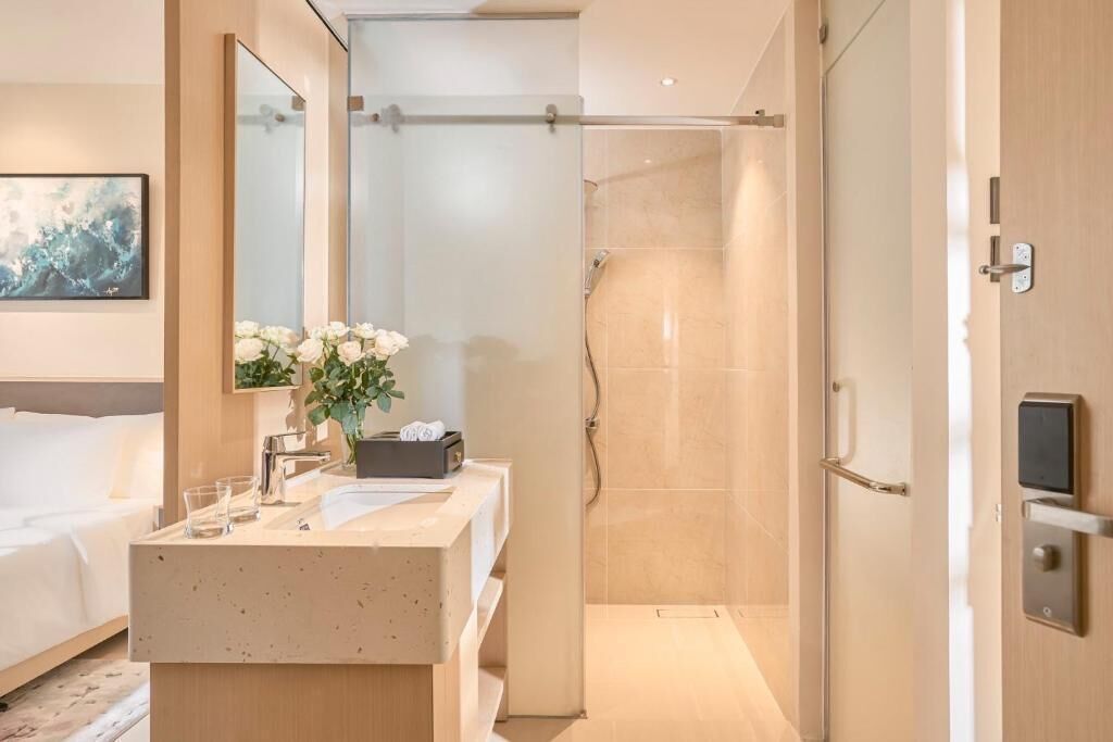Un baño de hotel moderno con ducha, lavamanos con espejo y mesita de noche con un jarrón de flores.