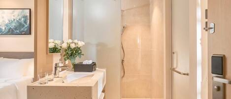 Un baño de hotel moderno con ducha, lavamanos con espejo y mesita de noche con un jarrón de flores.