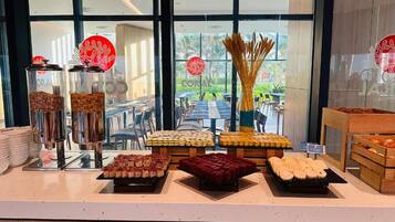 A buffet area with various food items displayed on trays and in containers, with a view of outdoor seating and greenery in the background.