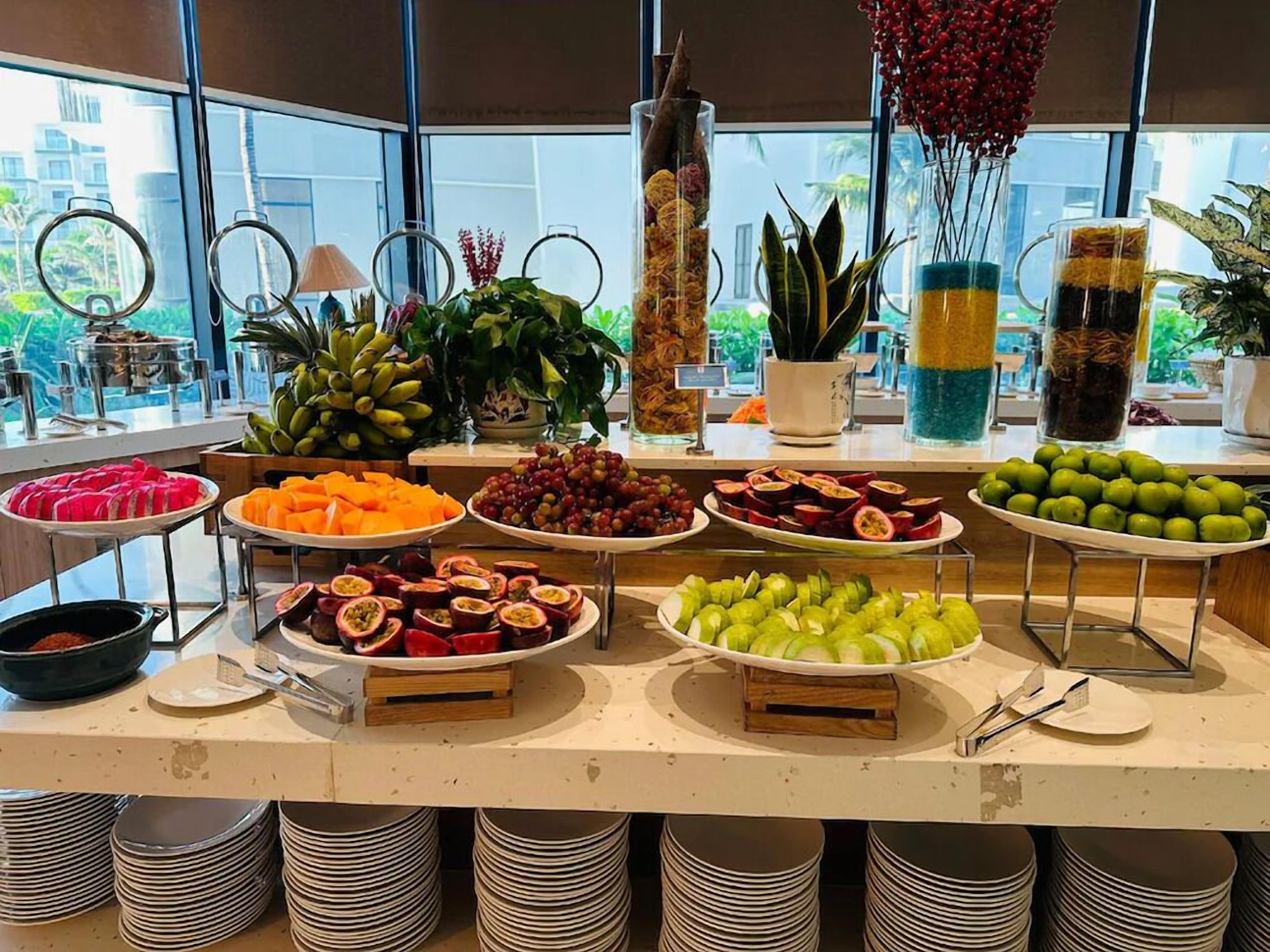 Un buffet proposant une variété de fruits et de légumes, notamment des bananes, des raisins et des pommes, disposés sur un comptoir d’où l’on peut admirer les bâtiments environnants.