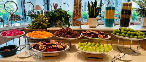 Un buffet proposant une variété de fruits et de légumes, notamment des bananes, des raisins et des pommes, disposés sur un comptoir d’où l’on peut admirer les bâtiments environnants.