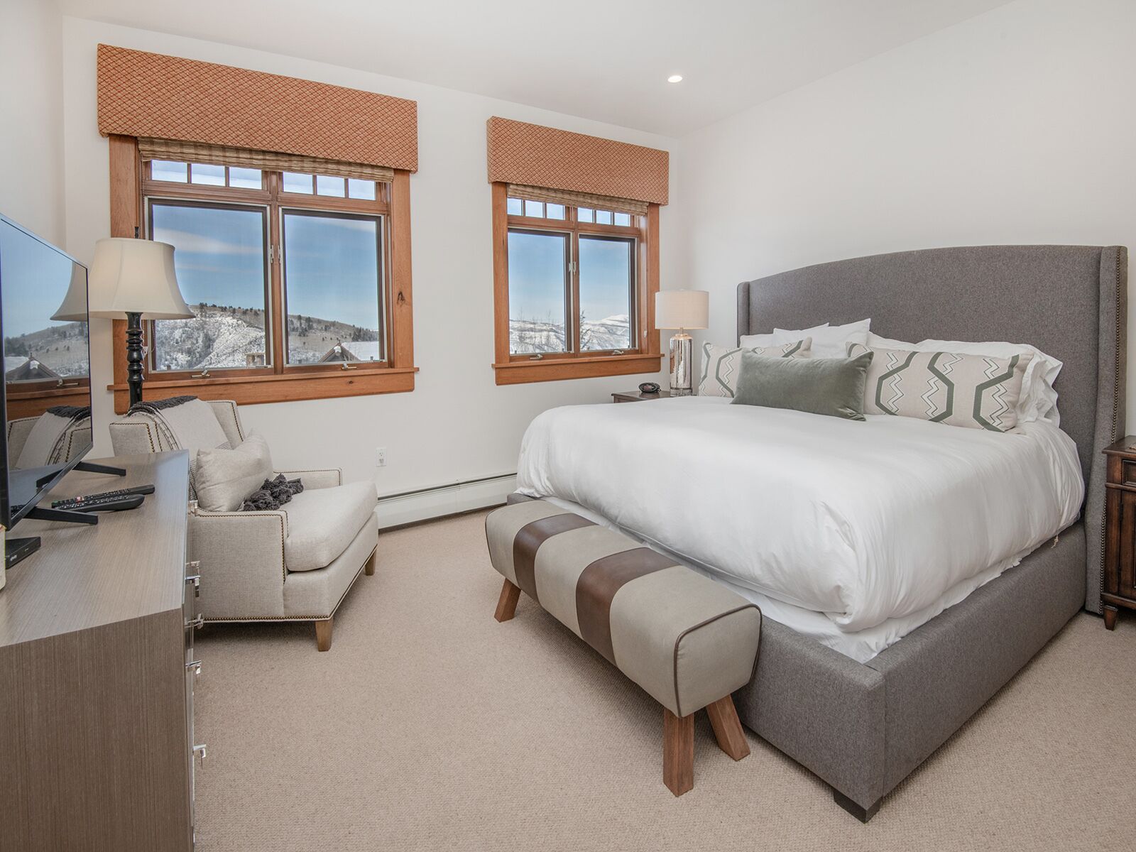 A bedroom with a large bed, two windows with blinds, a desk with a lamp, and a chair.